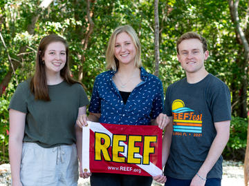 Three people in front of trees, holding a dive flag with the words "REEF" written on it in yellow.