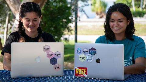 Two women, one in a black shirt and one in a teal shirt, sitting in front of laptops.