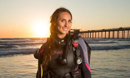 A dark-haired woman wearing scuba diving gear, smiling and standing in front of a sunset.